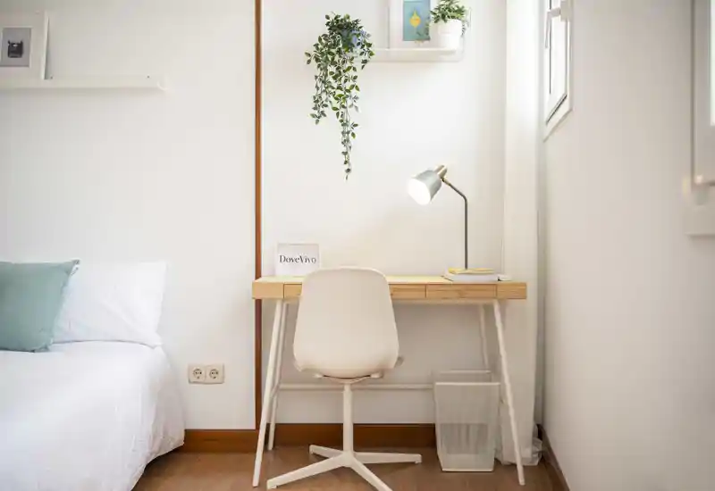 Rincón de dormitorio luminoso y minimalista con cama cómoda y un pequeño escritorio de madera junto a la ventana — ideal para descansar y trabajar de forma ligera.