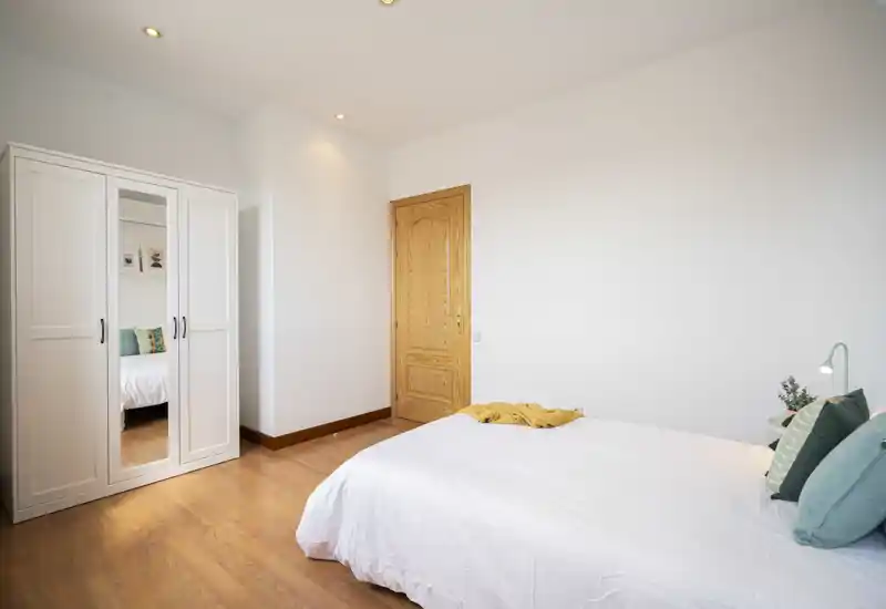 Dormitorio luminoso y minimalista con cama blanca, cojines turquesa y armario blanco con espejo. Estilo sencillo y moderno y suelos de madera cálidos que transmiten una atmósfera tranquila y luminosa, ideal para anuncios.