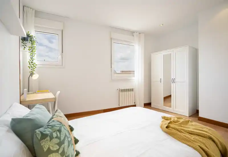 Dormitorio luminoso y minimalista con una amplia cama blanca, cojines verdes y una manta mostaza. Dos ventanas aportan luz natural abundante y un armario con espejo proporciona almacenamiento práctico.