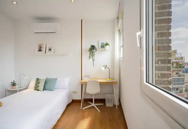 Chambre simple lumineuse et chaleureuse avec un lit confortable, un bureau en bois et une grande fenêtre apportant beaucoup de lumière naturelle.