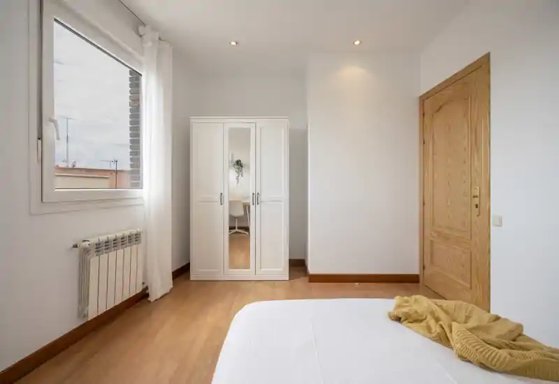 Rincón de dormitorio luminoso y de mobiliario sencillo con un armario blanco, puerta de madera y la cama parcialmente visible en primer plano; la luz natural de la ventana genera una sensación aireada.