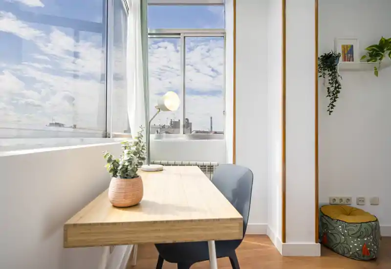Coin salon moderne et lumineux avec un bureau en bois près de grandes fenêtres, une chaise confortable, une plante en pot et une lampe, offrant un espace de travail ensoleillé et accueillant.