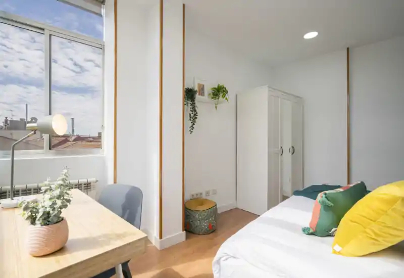 Chambre simple moderne et lumineuse avec lit bien fait, coussins colorés, armoire et un coin bureau près d'une grande fenêtre baigné de lumière naturelle.