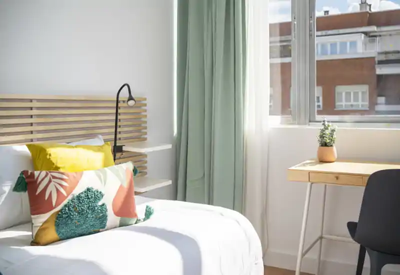 Chambre lumineuse et élégante avec un lit blanc confortable, des coussins décoratifs et une tête de lit en bois ; lumière naturelle abondante depuis une grande fenêtre près d'un bureau.