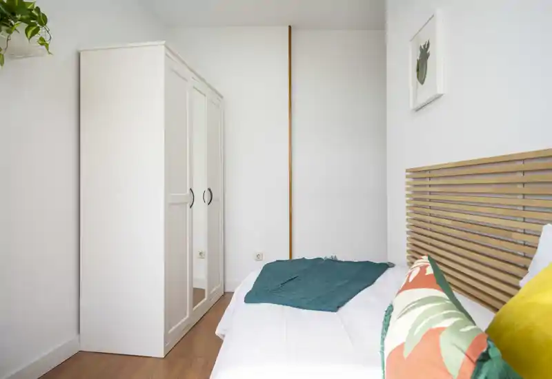 Chambre lumineuse et minimaliste avec une tête de lit en lattes de bois, une armoire blanche avec miroir et des coussins colorés pour une touche conviviale.