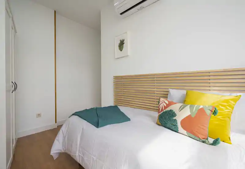 Chambre compacte et lumineuse, bien mise en scène avec une tête de lit à lattes en bois, coussins colorés et couette blanche. Sol imitation bois, placard compact et climatisation au-dessus du lit.