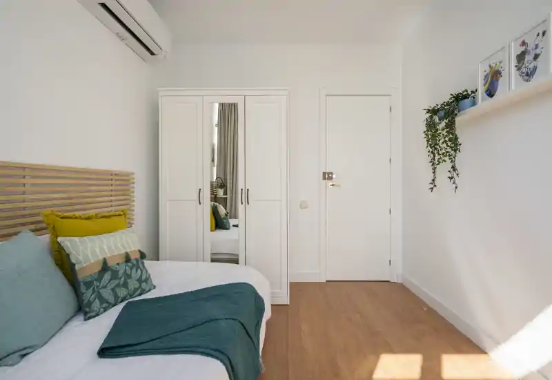 Chambre moderne et lumineuse avec une tête de lit en bois, coussins bleu-vert et jaune, une armoire blanche avec miroir et un sol en bois chaleureux.