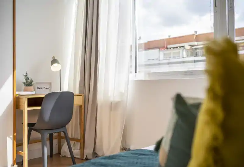 Coin chambre lumineux et moderne avec un lit bien fait au premier plan, un petit bureau en bois et une chaise près d'une fenêtre ensoleillée — parfait pour le repos et le télétravail.