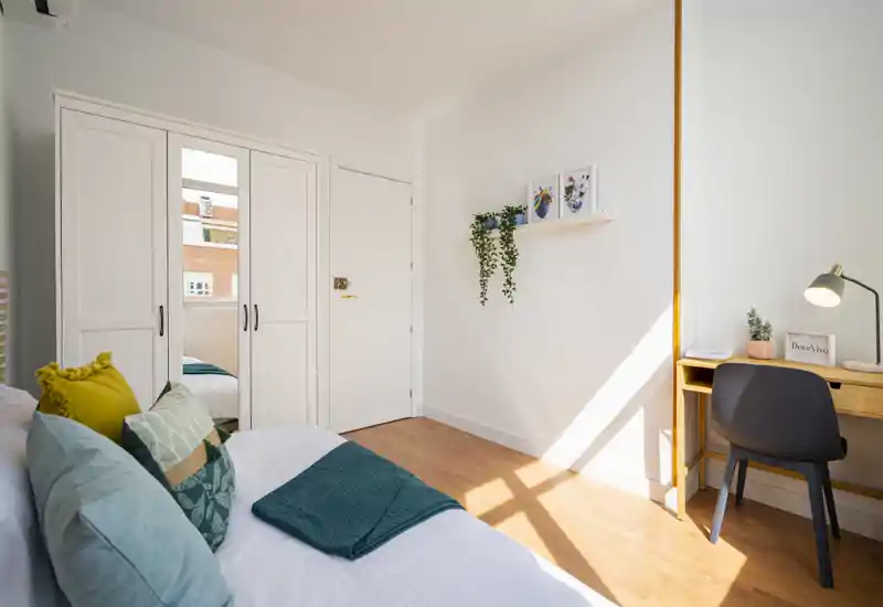 Bright, styled bedroom with a comfortable double bed, decorative cushions and a small wooden desk by the window letting in warm sunlight.