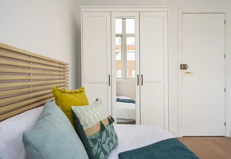 Chambre lumineuse et moderne avec tête de lit en lamelles de bois, coussins décoratifs bleu-vert et jaune superposés, et une armoire blanche avec miroir reflétant la fenêtre — espace de nuit frais et accueillant.