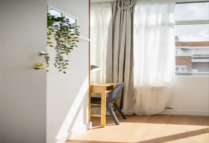 Espace de vie lumineux et minimaliste avec un petit bureau en bois, une chaise moderne et des rideaux doux; la lumière du jour inonde la pièce pour une atmosphère calme.