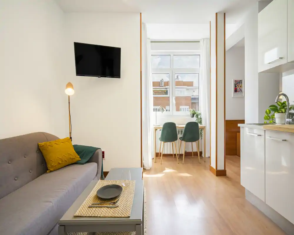 Area living luminosa e accogliente con divano comodo, TV a parete e un piccolo angolo pranzo davanti alla finestra — perfetta per un appartamento compatto.