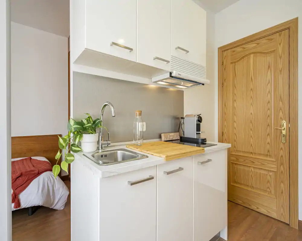 Angolo cottura compatto e luminoso con mobili bianchi, lavello in acciaio, piano di legno e macchinetta del caffè. L’ambiente è arredato con una pianta e una porta in legno caldo, perfetto per un monolocale.