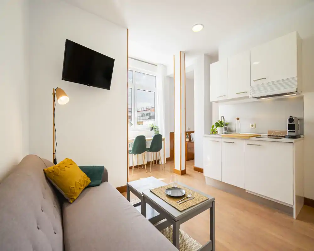 Zona giorno luminosa e moderna con angolo cottura compatto, divano con cuscini, TV a parete e un piccolo tavolo vicino alla finestra — perfetta per affitti brevi.