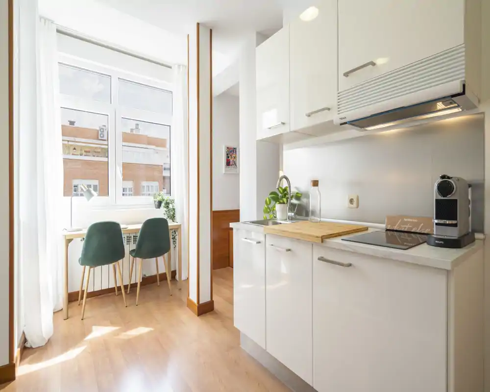 Cucina moderna compatta con mobili bianchi, piano cottura a induzione e macchinetta per caffè sul piano; ampia finestra che illumina l'ambiente e un angolo colazione/ufficio con due sedie verdi.