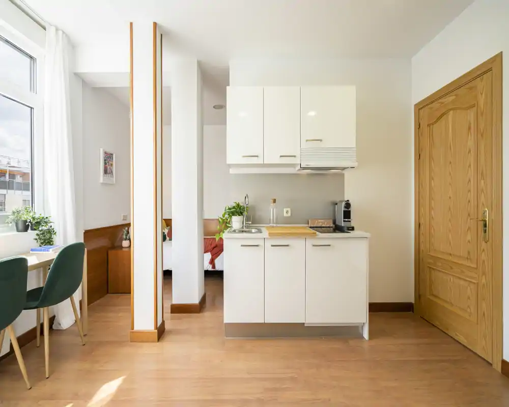Angolo stanza luminoso in stile monolocale con angolo cottura compatto e una nicchia letto visibile; la luce naturale illumina lo spazio e il pavimento in legno dona calore.