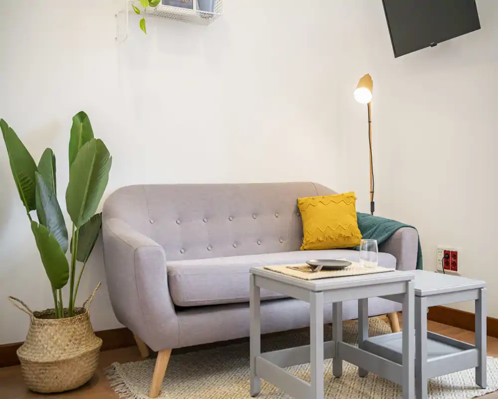 Accogliente angolo living con divano grigio chiaro, tappeto testurizzato, tavolini annidati e lampada da terra: ambiente curato e ideale per rilassarsi.