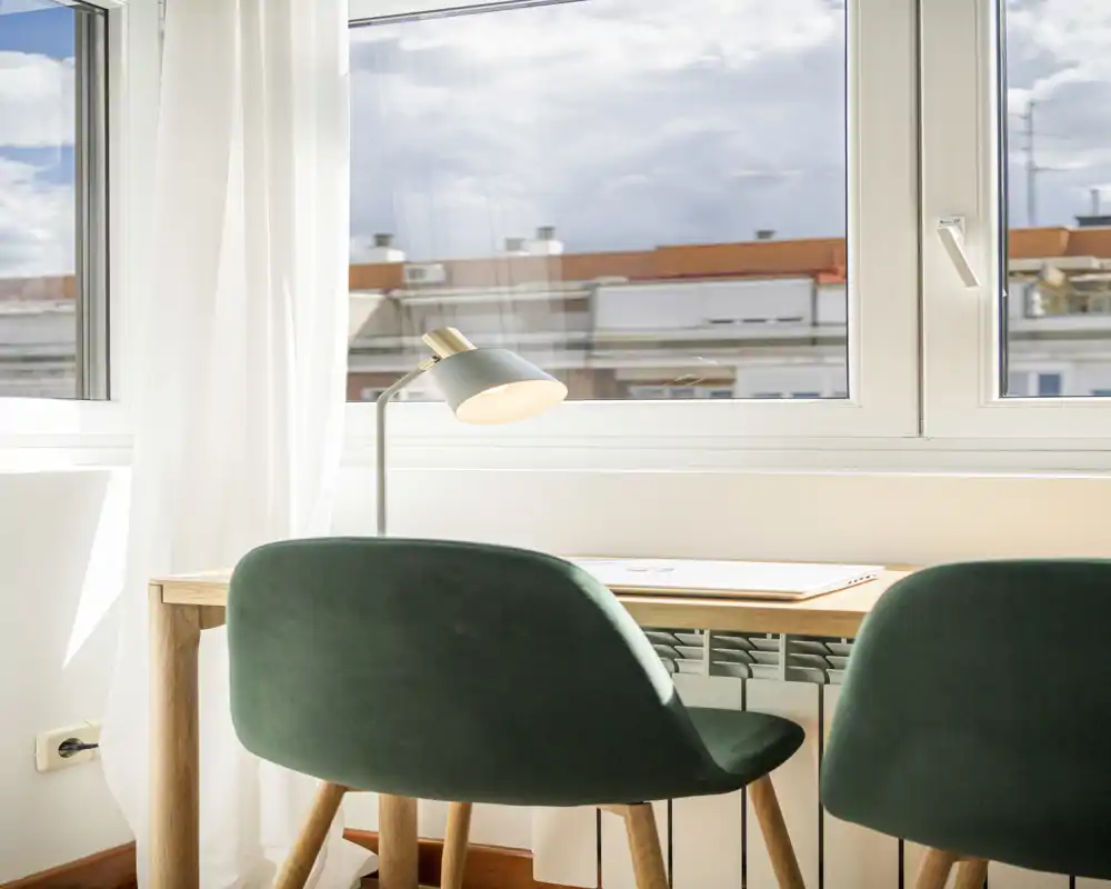 Angolo lavoro luminoso vicino a una grande finestra con due sedie imbottite e una scrivania in legno semplice, ideale per smart working o lettura.