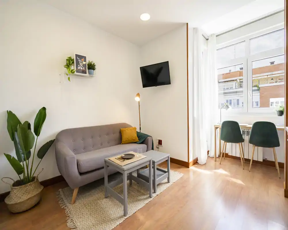 Zona giorno luminosa e ben arredata con divano compatto, tavolini e una postazione vicino alla finestra — perfetta per rilassarsi o lavorare da casa.