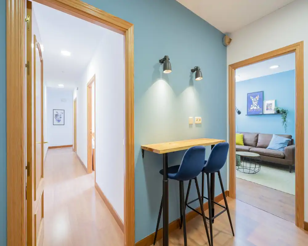 Bright, modern living area seen through a doorway featuring a comfortable sofa, cushions and a decorative shelf; a small bar-style table and stools in the foreground add a casual dining/work corner.