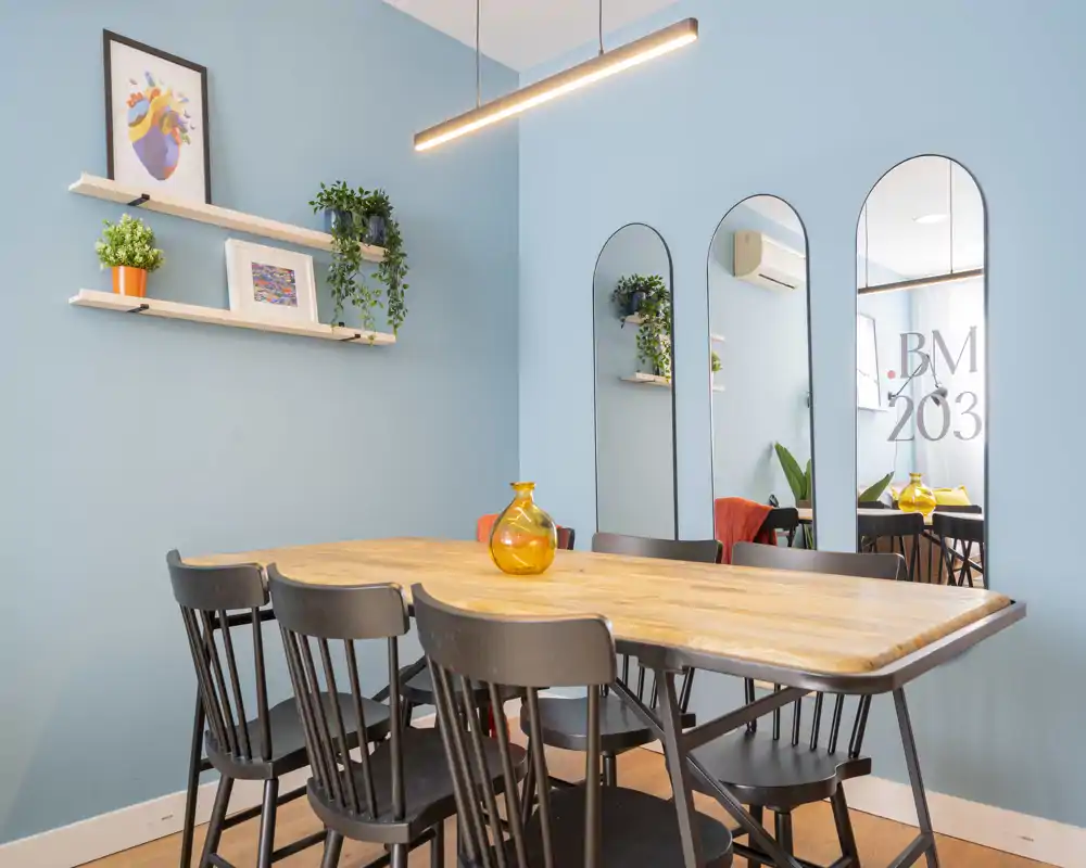 Bright, styled dining area with a wooden table, six black chairs and decorative mirrors on a soft blue wall — ideal for shared meals and small gatherings.