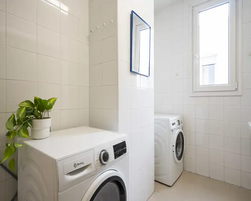 Bright, tiled laundry/utility room with two front-loading washing machines, a small window and a potted plant on top of one machine. Clean, well-lit space ideal for appliance-focused listings.