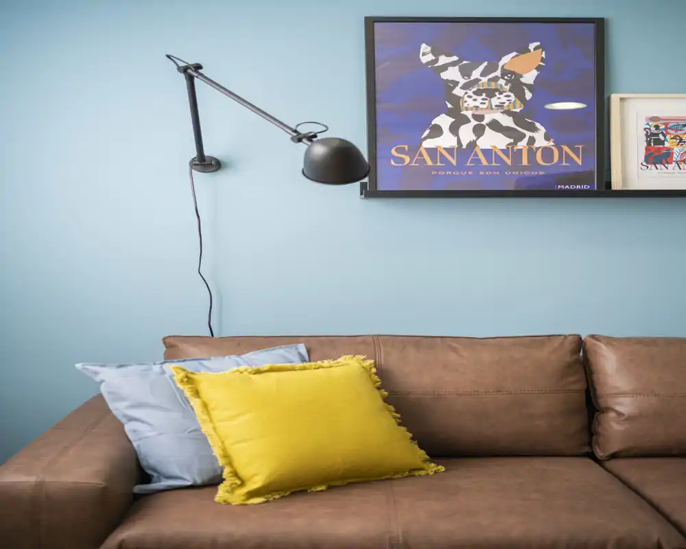 Stylish living area with a brown leather sofa, colorful cushions and framed art on a soft blue wall — bright, cozy and well-styled for a rental listing.