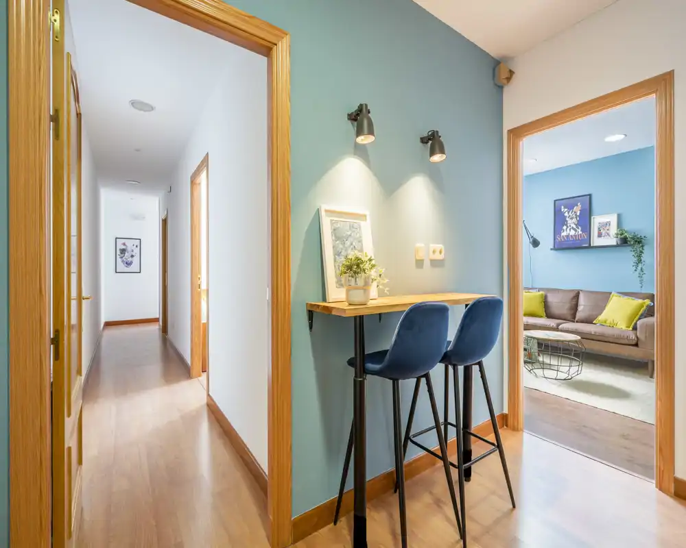 Bright, modern entrance/hallway with a slim bar table and two stools against a teal accent wall, leading to a living room and corridor.