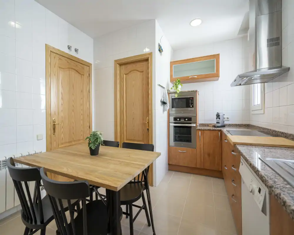 Bright, modern kitchen with wooden cabinets, built-in oven and a wooden dining table for four. Clean tiled walls and granite countertops create a neat, functional space.