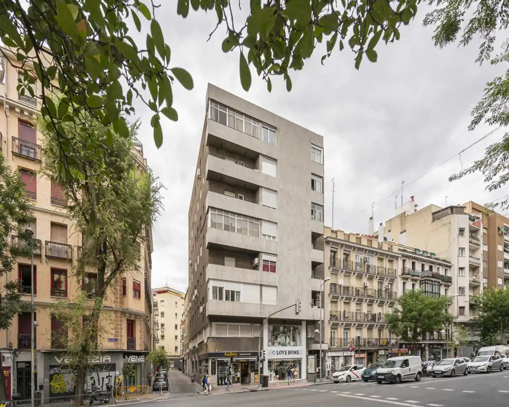 Street view of an urban building facade with shops at ground level and several balconies; framed by trees and neighboring buildings under an overcast sky.