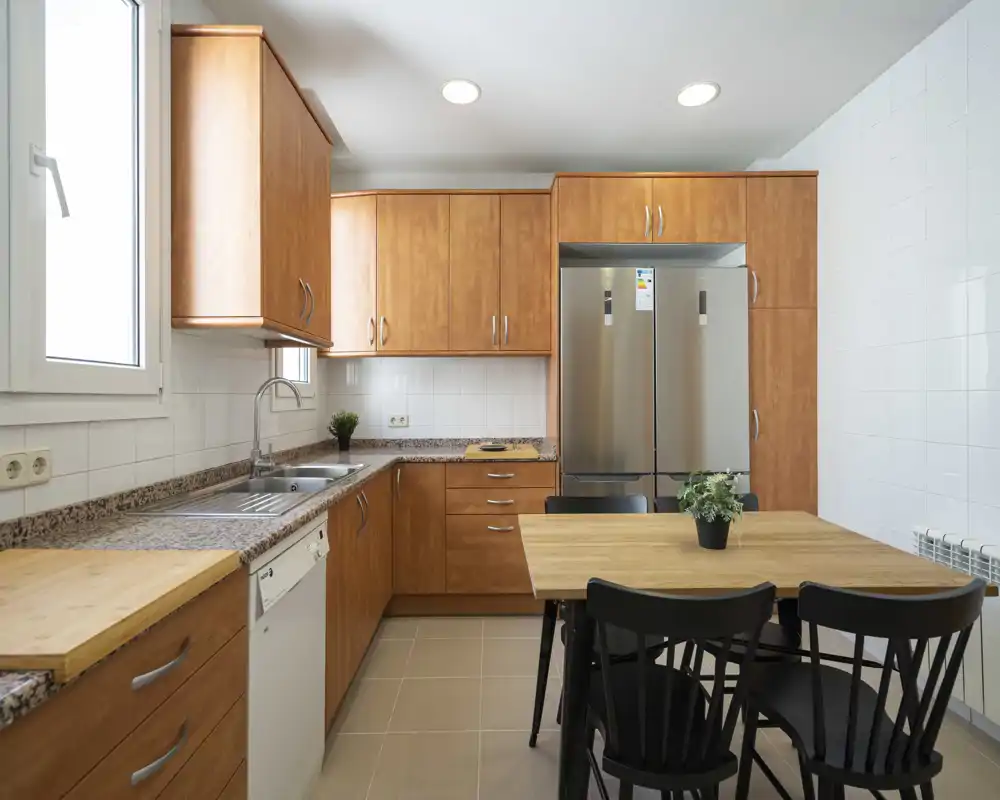 Bright modern kitchen with warm wooden cabinets, granite countertops, a stainless-steel double-door refrigerator and a dining table with chairs — clean, functional space ideal for everyday use.