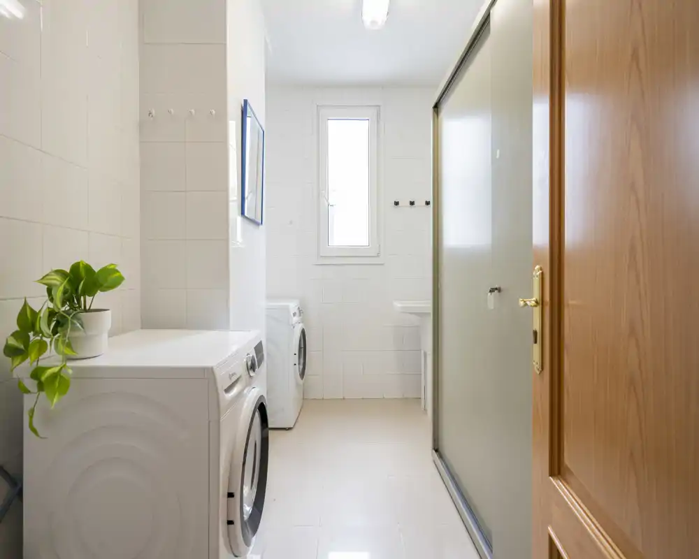 Bright, tiled utility room with two front-loading washing machines, a small sink and a frosted glass shower/partition. The space is clean and functional, with natural light from a narrow window and a potted plant adding a touch of greenery.