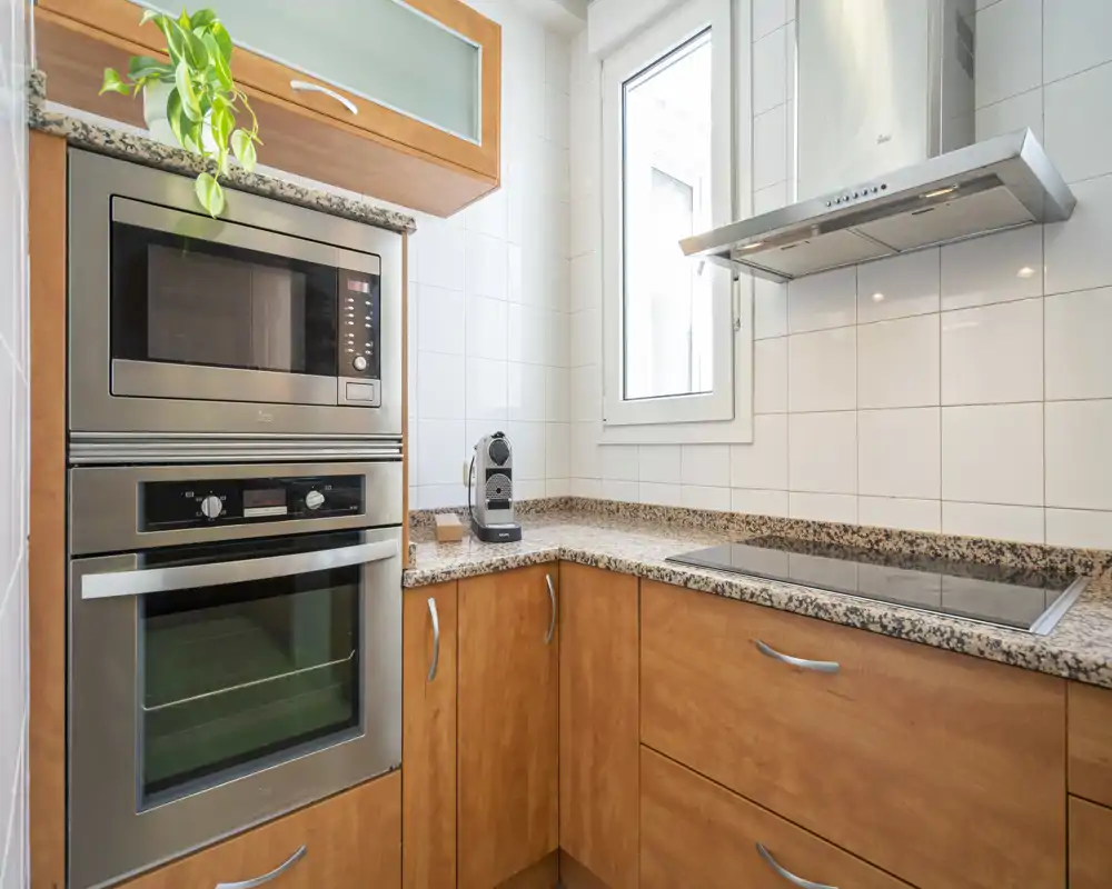 Compact, modern kitchen with built-in stainless-steel oven and microwave, granite countertops, glass-ceramic hob and natural light from a window.