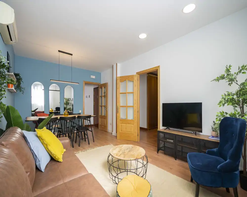 Bright, stylish living room with a comfortable leather sofa, accent chair, TV unit and a dining area in the background; modern decor with plants and coordinated cushions.
