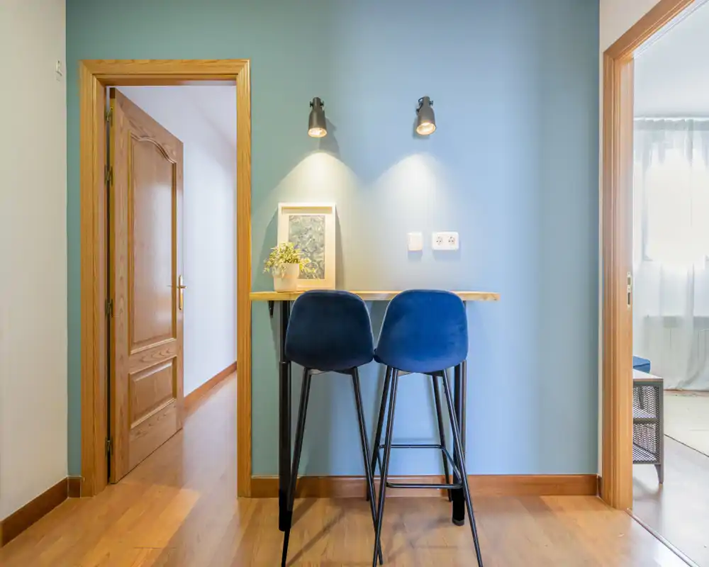 Bright, compact living area with a small bar-style table and two blue stools against a soft blue wall, wooden flooring and warm spot lighting creating a cozy nook.