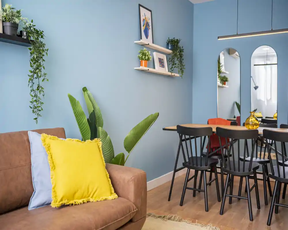 Stylish and cozy living/dining area with a brown sofa accented by colorful cushions, indoor plants and a sleek dining table with black chairs — bright, modern and inviting.