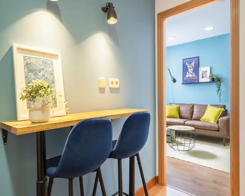 Bright, stylish living area seen through a doorway with a cozy sofa, colorful cushions and a small coffee table; foreground shows a compact wooden bar desk with two blue stools and decorative lighting.