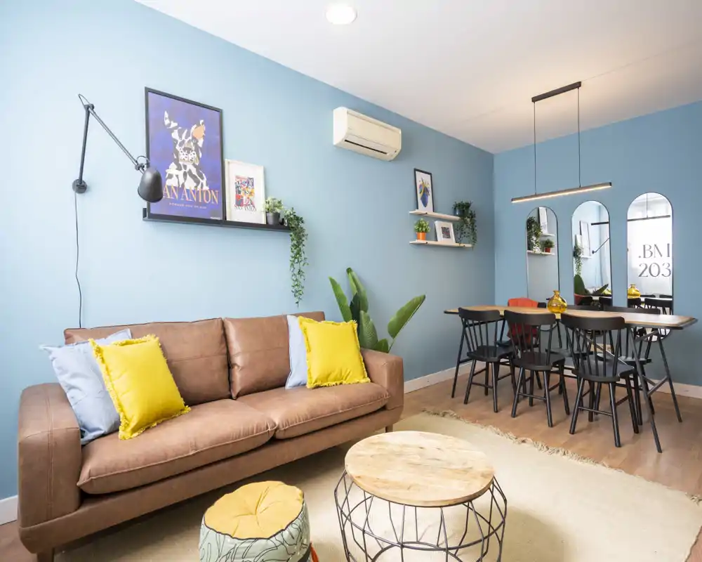 Bright, stylish living room with a brown sofa, colorful cushions, decorative plants and a dining area in the background — ideal for comfortable socializing and modern rental listings.