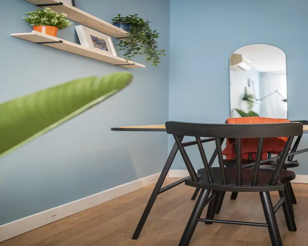 Bright, styled dining area with a wooden table and black chairs, leafy plants and decorative shelves against a soft blue wall — a cozy spot for meals.