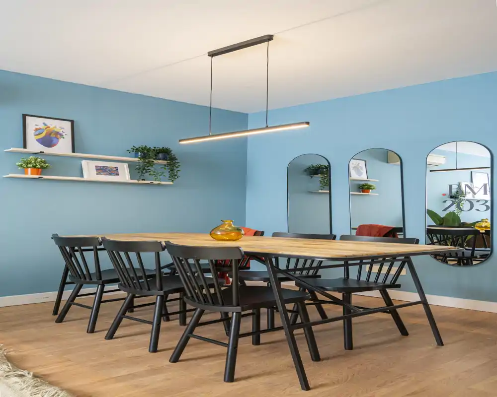 Bright, modern dining room with a long wooden table, black chairs and blue accent walls; stylish pendant light and decorative mirrors enhance the space.