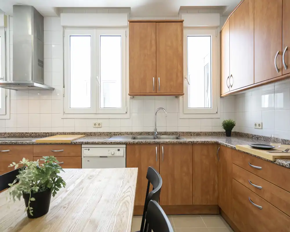 Bright, modern kitchen with wooden cabinets, granite countertops and a dining table in the foreground. Natural light from the windows creates a warm, inviting atmosphere.