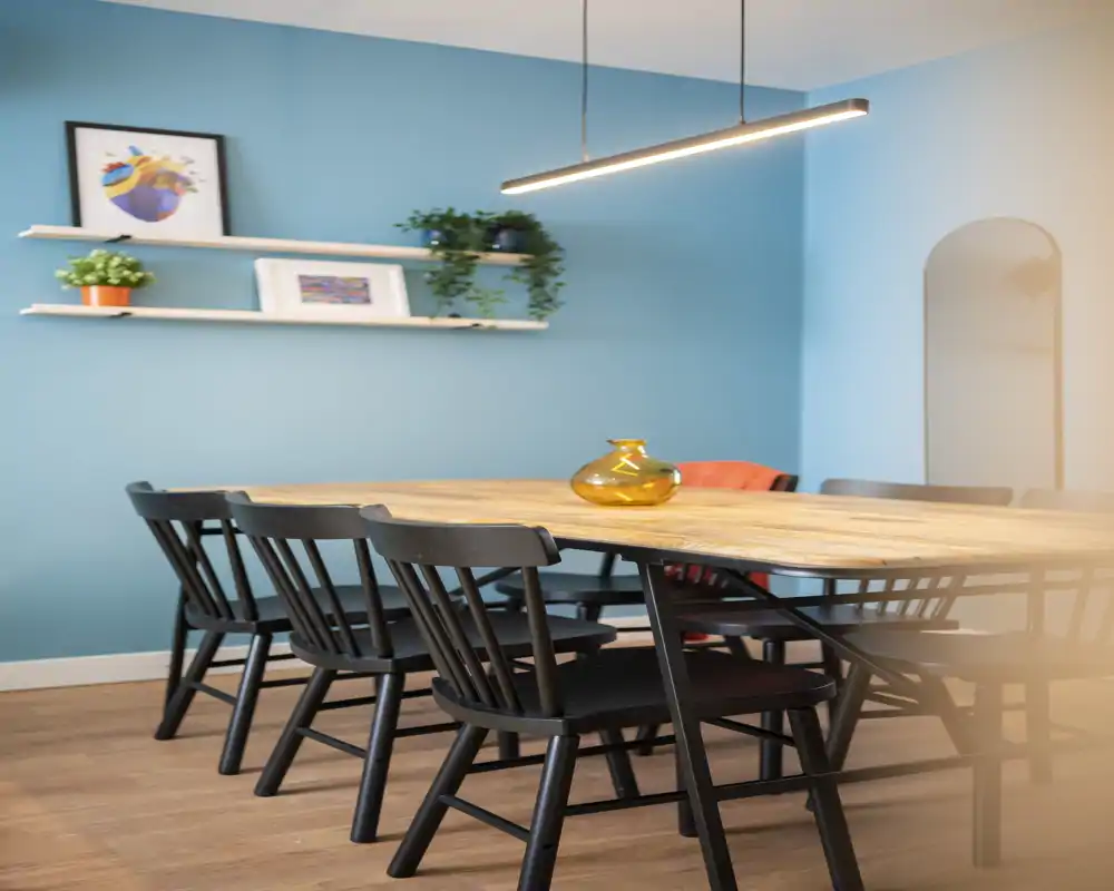 Bright, modern dining room with a long wooden table and several black chairs; decorative shelving and pendant lighting create a welcoming dining space.