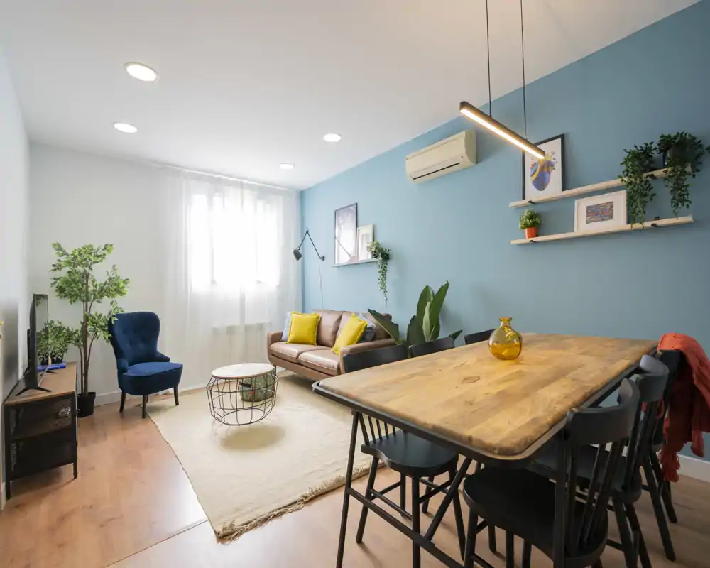 Bright, stylish living room with a comfortable sofa, accent armchair, indoor plants and a wooden dining table — well-lit by a large window and soft ceiling lights.