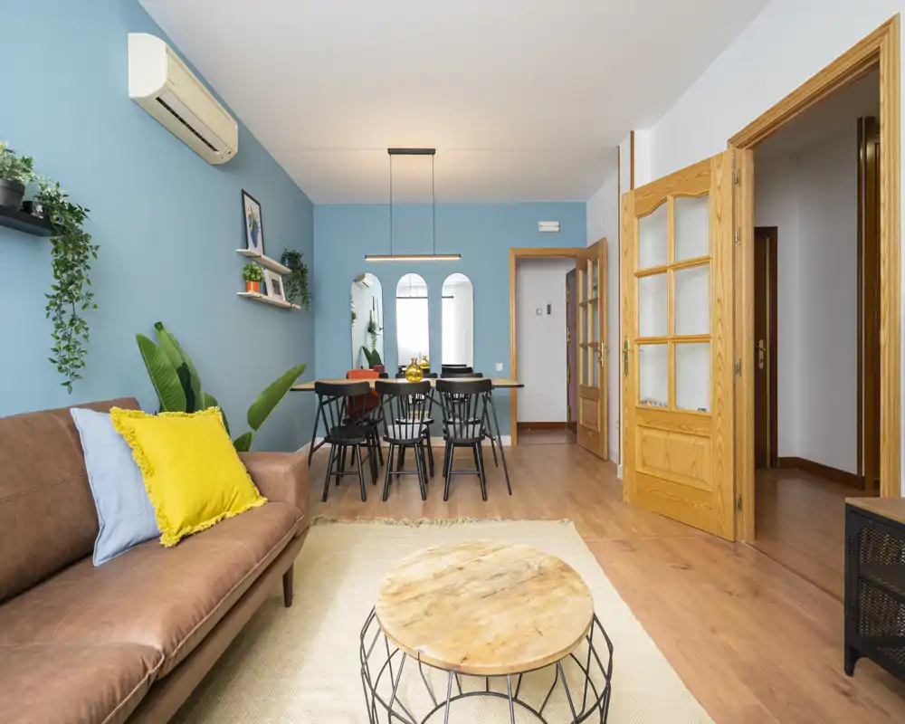 Bright, stylish living and dining area featuring a leather sofa with colorful cushions, a round wooden coffee table, and a dining table for six against a soothing blue accent wall.