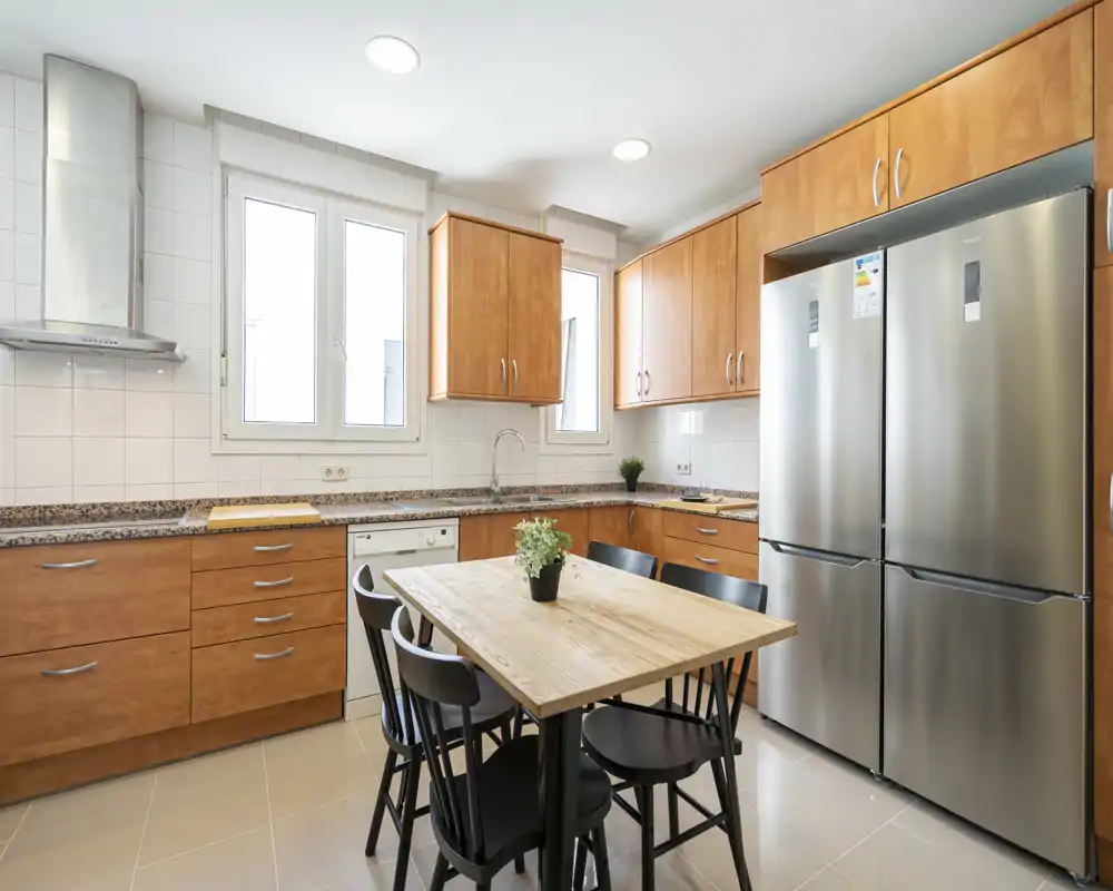 Bright modern kitchen with wooden cabinets, granite countertops, a central dining table and stainless steel fridge — ideal for family meals and social cooking.