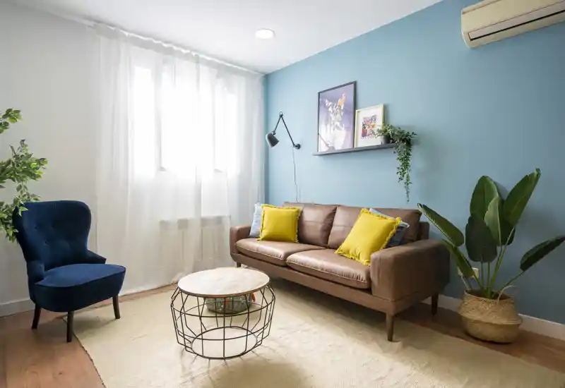 Salón luminoso y con estilo con sofá de cuero, cojines coloridos, sillón de acento y plantas de interior; la luz natural a través de las cortinas transparentes genera un ambiente acogedor.