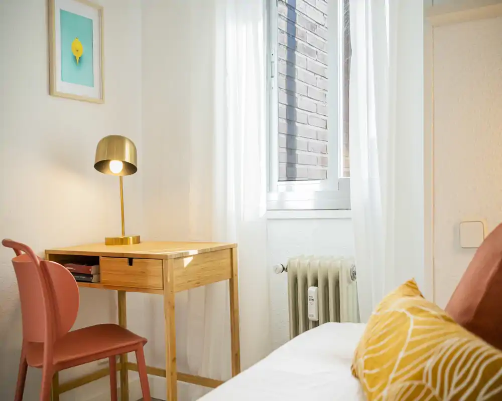 Angolo camera luminoso e accogliente con una piccola scrivania in legno, lampada moderna e letto con cuscini decorativi — perfetto per riposare e leggere o lavorare.
