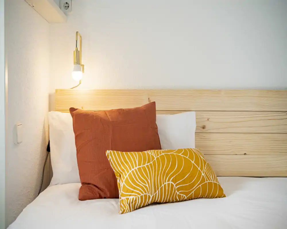 Angolo di camera luminoso e accogliente con testiera in legno, biancheria bianca e due cuscini decorativi dai toni caldi. Una lampada a parete crea un’atmosfera invitante, ideale per foto di annunci.