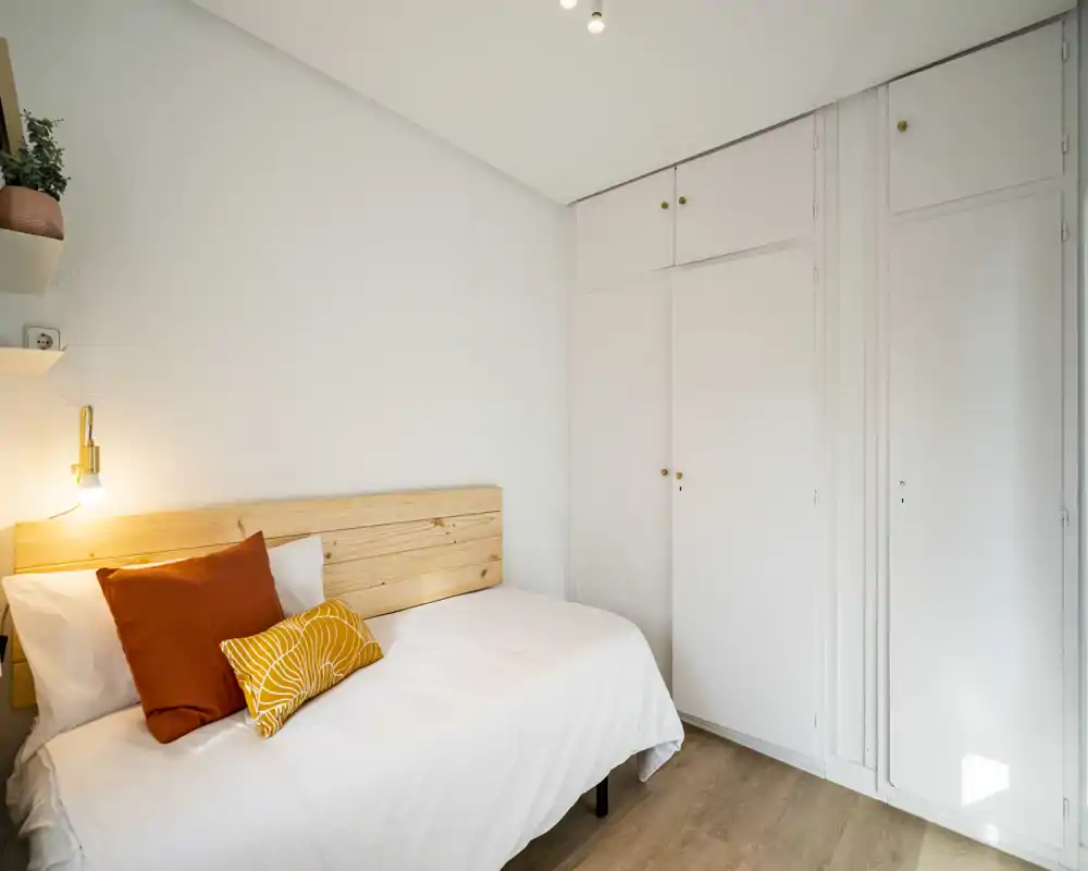 Camera da letto luminosa e compatta con letto singolo, testiera in legno chiaro e cuscini decorativi; pareti bianche e armadio a muro sfruttano lo spazio.
