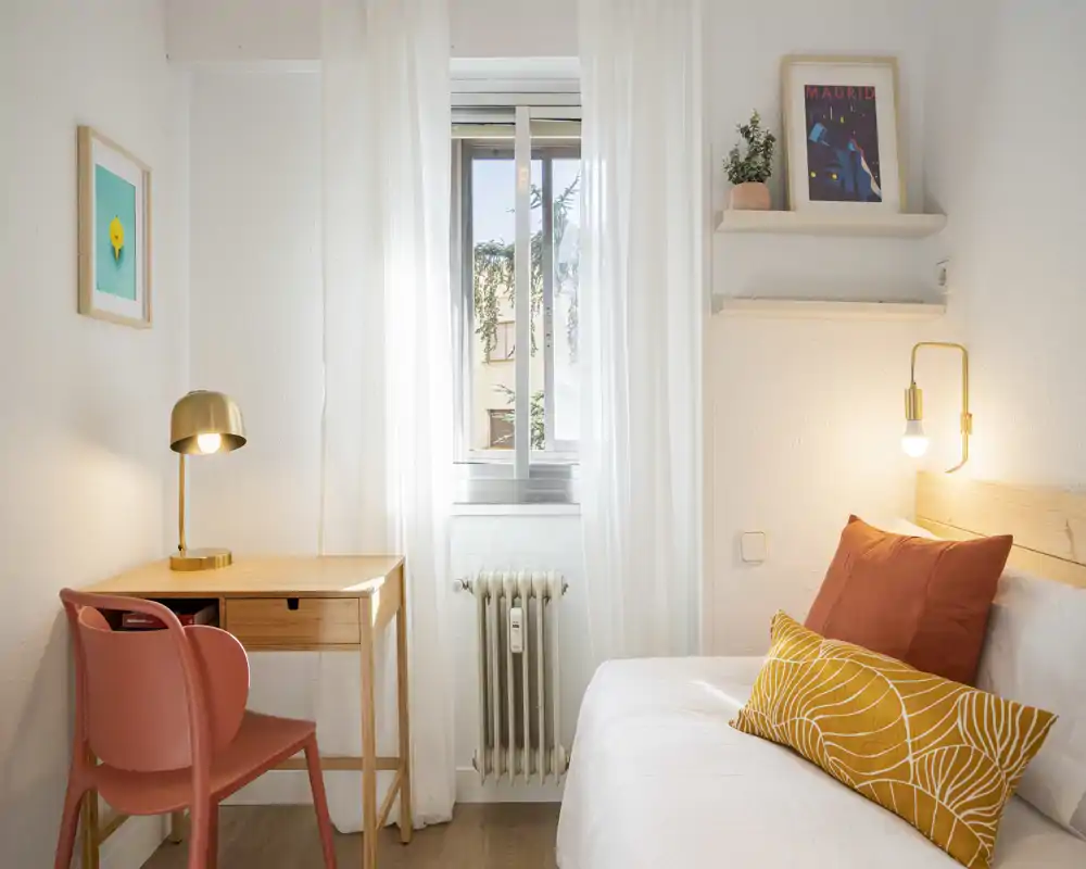 Camera da letto luminosa e accogliente con letto ben sistemato, cuscini decorativi e luce calda al comodino. Una piccola scrivania e una sedia vicino alla finestra rendono lo spazio compatto e funzionale.